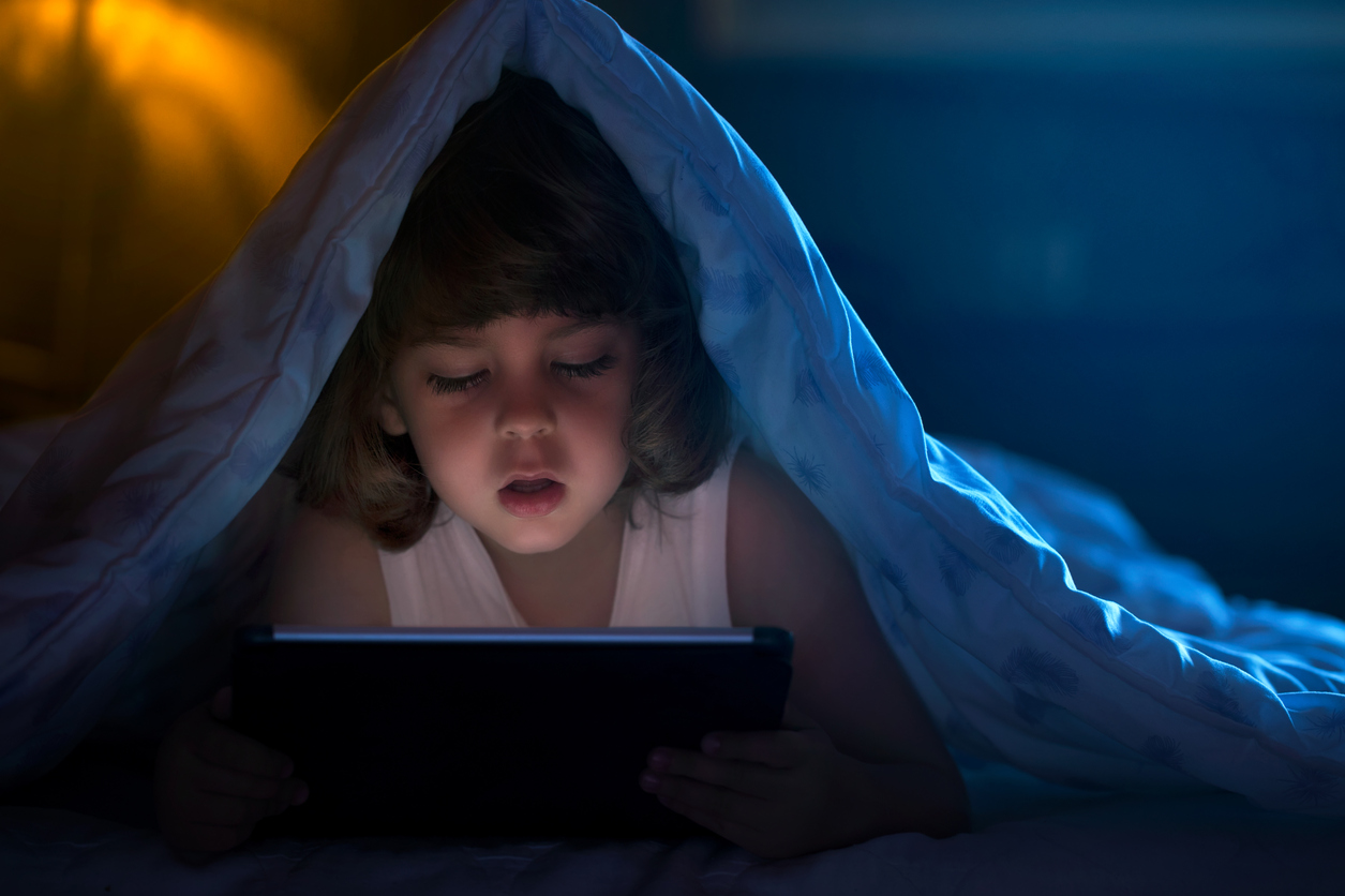 copil in pat sub patura uitandu-se pe tableta