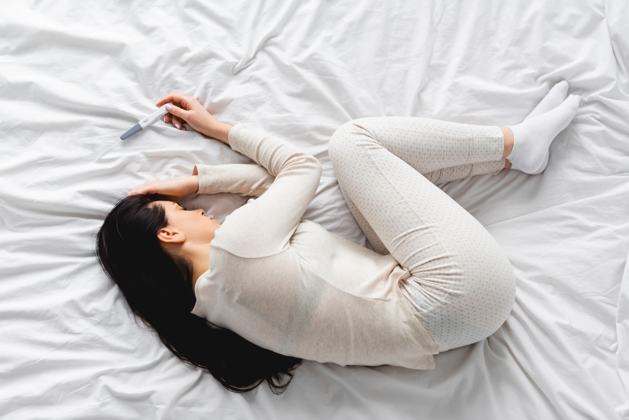 femeie care sta ghemuita in pat cu un test de sarcina negativ in mana