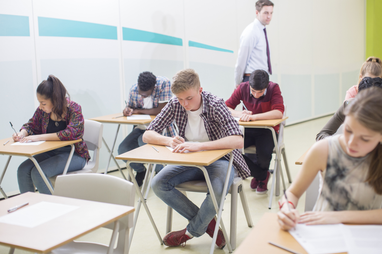 elevi care sustin un examen asezati in banci scriind si supravegheati de un profesor