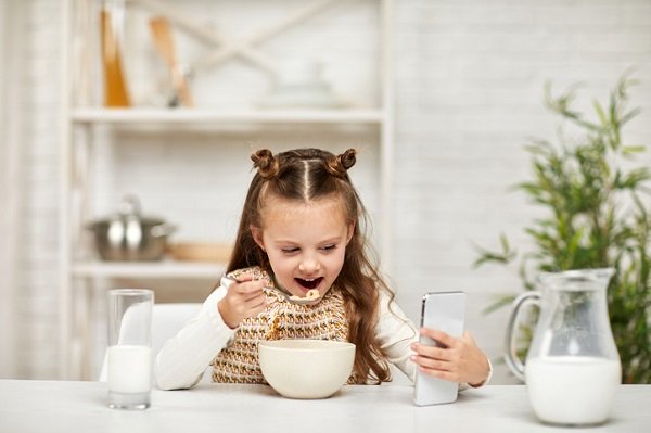 fetita mananca cereale cu lapte privind in telefon