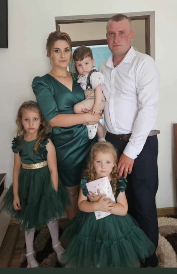 barbat in camasa alba langa femeie imbracata in verde care tine un copil in brate si doua fete imbracate la fel langa ei