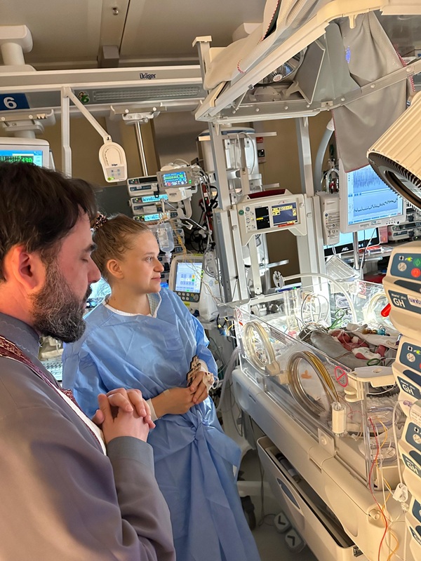 femeie in halat albastru alaturi de un preot in sectia de neonatologie