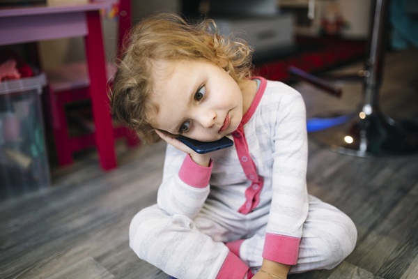 fetita imbracata in pijamale cu bucle blonde sta pe jos intr-o incapere si tine un telefon mobil la ureche