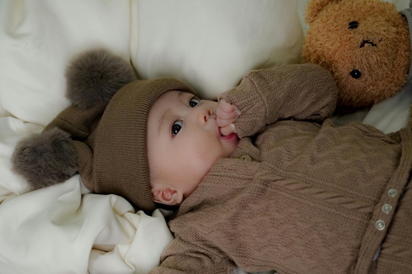 bebelus stand pe spate in pat imbracat intr-un costum gros maro cu o caciula maro pe cap isi suge degetul