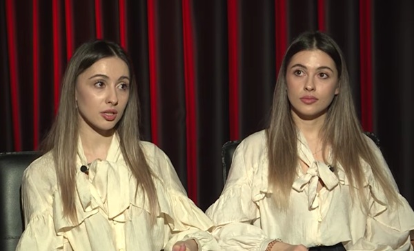 doua surori gemene imbracate la fel cu parul lung blond pe fundalul unei copertine grena