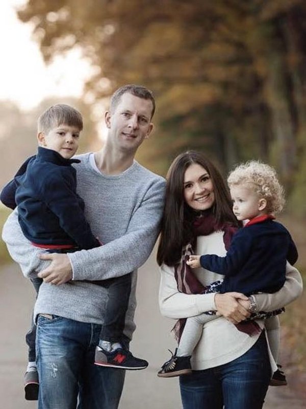 o mama cu parul lung brunet tine un copil in brate langa un tata care tine celalalt copil in brate pe strada