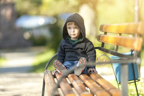 copil imbracat in hanorac negru sta pe o banca in parc singur