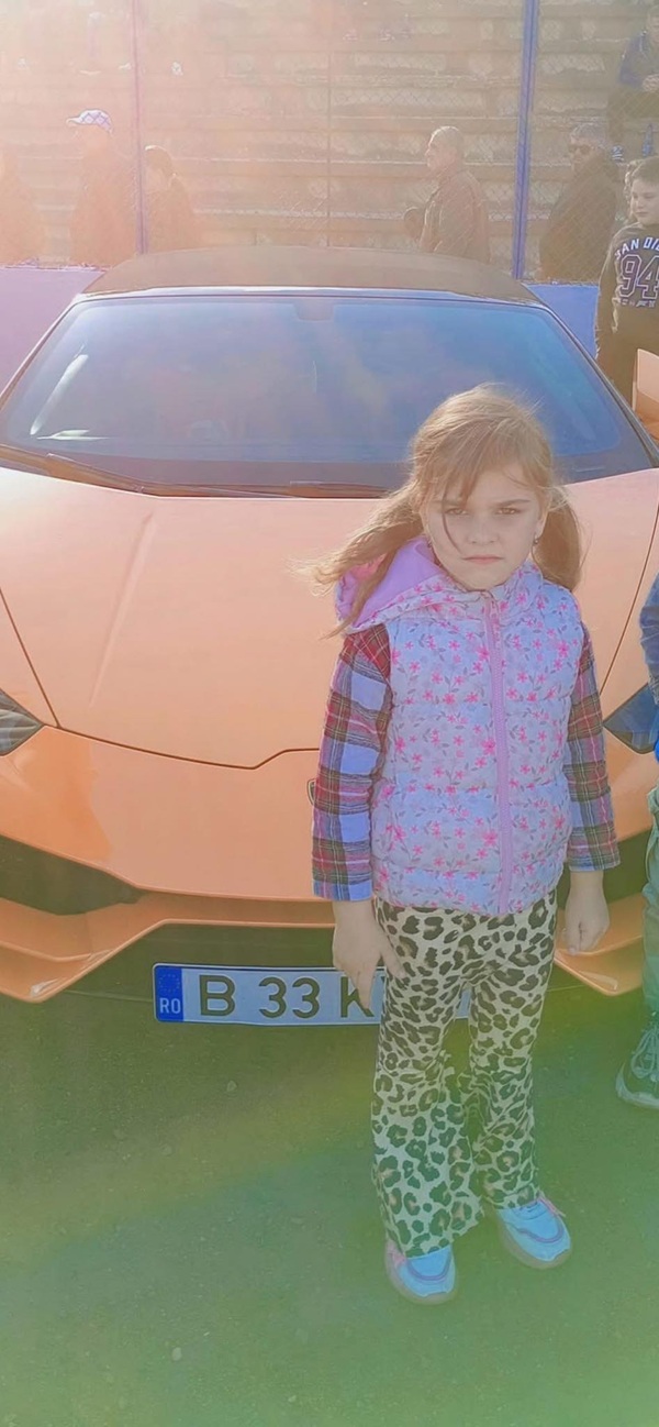 o fetita cu parul lung si vesta mov in fata unei masini rosii