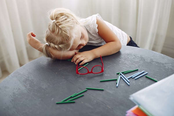 fetita blonda cu parul prins sta cu capul pe masa langa creioane colorate