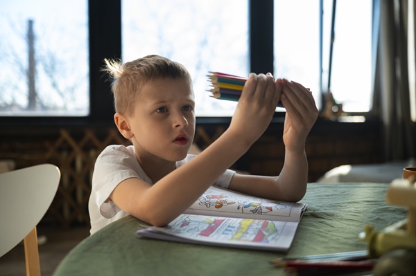 copil blond sta la masa cu un caiet in fata se uita la creioane colorate pe care le tine in mana