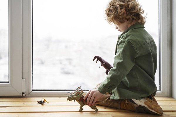 un baiat blond cret se joaca cu dinozauri pe marginea unui geam la etaj