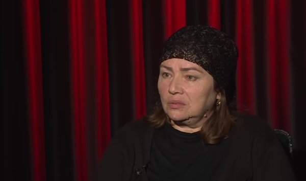 femeie cu parul saten imbracata in negru cu batic negru pe cap da interviu pe fundal o copertina grena