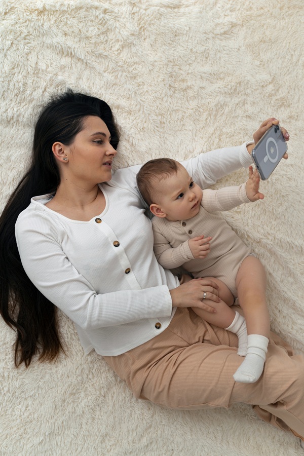 mama cu parul lung negru imbracata in bluza alba si pantaloni crem face un selfie cu bebelusul ei in pat