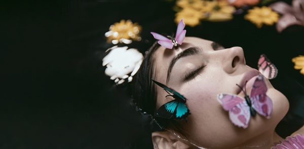 femeie cu fluturi colorati si cu flori pe fata sta cu fata in sus si capul in apa