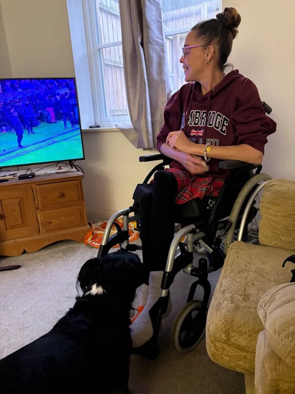 femeie in scaun cu rotile se uita la tv cu un caine langa ea
