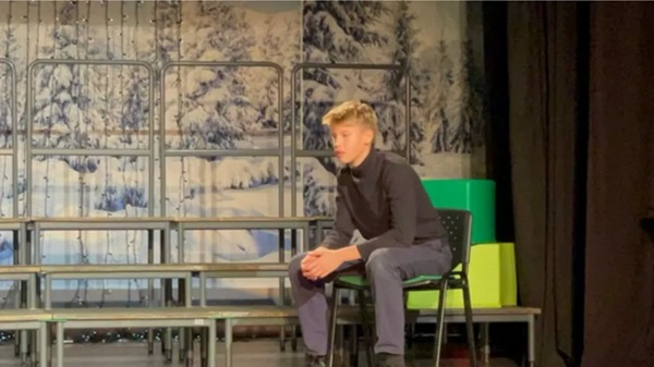 un baiat cu parul blond sta pe un scaun pe scena