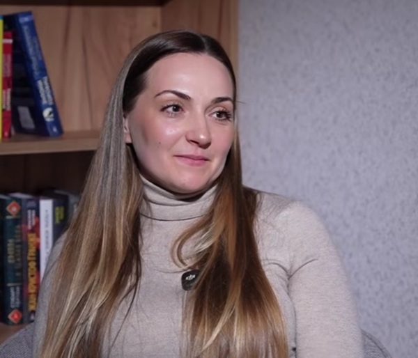 femeie cu parul lung blond in bluza gri in spate o biblioteca cu carti