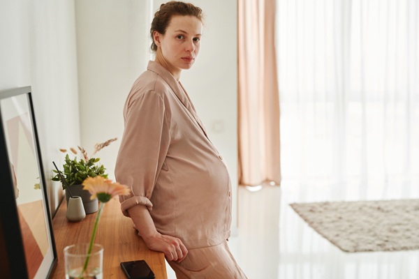 femeie gravida imbracata in rochie roz se sprijina de un dulap