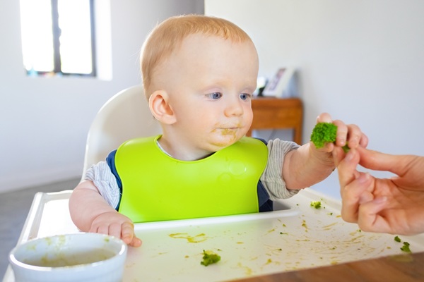 copil cu bavetica verde sta la masa si mananca broccoli