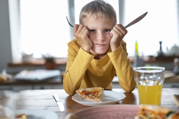 copil imbracat in galben tine furculita si cutit in mana la masa in fata are o felie de pizza