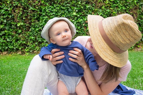 mama cu palarie in cap tine bebelus imbracat in albastru afara in soare
