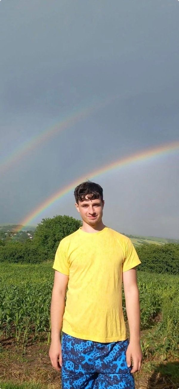 baiat cu tricou galben si pantaloni albastri pe un camp in spate un curcubeu