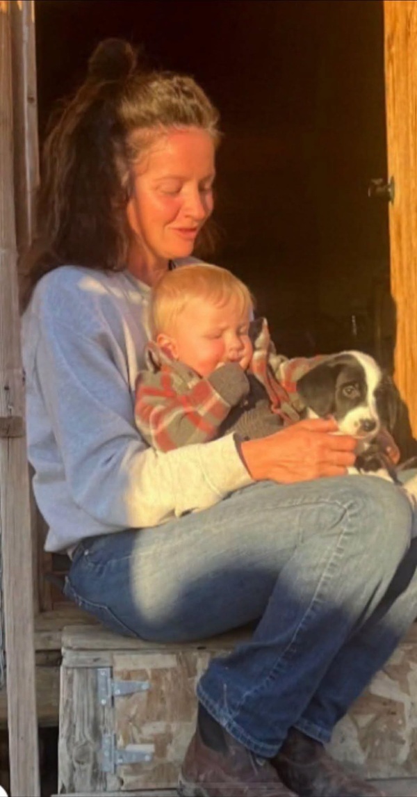 mama imbracata in blugi si camasa bleu isi tine copilul in brate pe veranda langa un caine