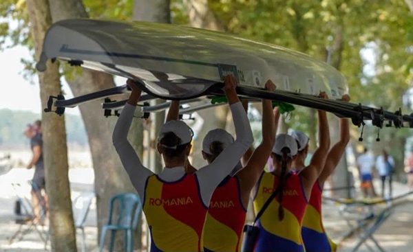 4 sportive romance intoarse cu spatele duc cu mainile in aer o barca de canotaj