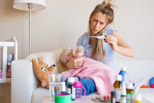 mama-blonda-care-vorbeste-la-telefon-si-este-agitata-si-care-tine-in-mana-un-termometru-si-tine-in-brate-o-fetita-blonda-care-bea-lapte-si-este-invelita-cu-o-patura-roz-iar-in-fata-ei-este-o-masa-plina-cu-obiecte-pentru-copii