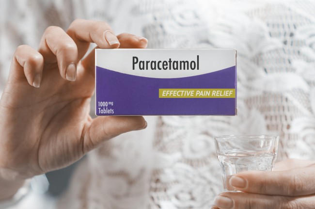 femeie-care-tine-intr-o-mana-o-cutie-de-paracetamol-cu-ambalaj-alb-si-mov-iar-in-cealalta-mana-are-un-pahar-cu-apa