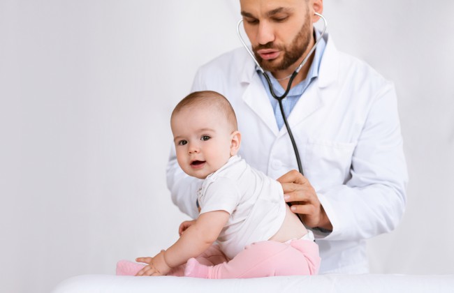 bebelusi-cu-pantaloni-roz-sta-si-se-uita-mirat-cat-timp-este-consultat-cu-stetoscopul-pe-spate-de-un-doctor