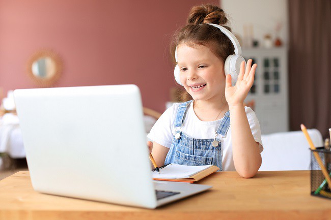 fetita-blonda-cu-salopeta-de-blugi-si-casti-albe-pe-cap-face-cu-mana-in-fata-unui-laptop-alb